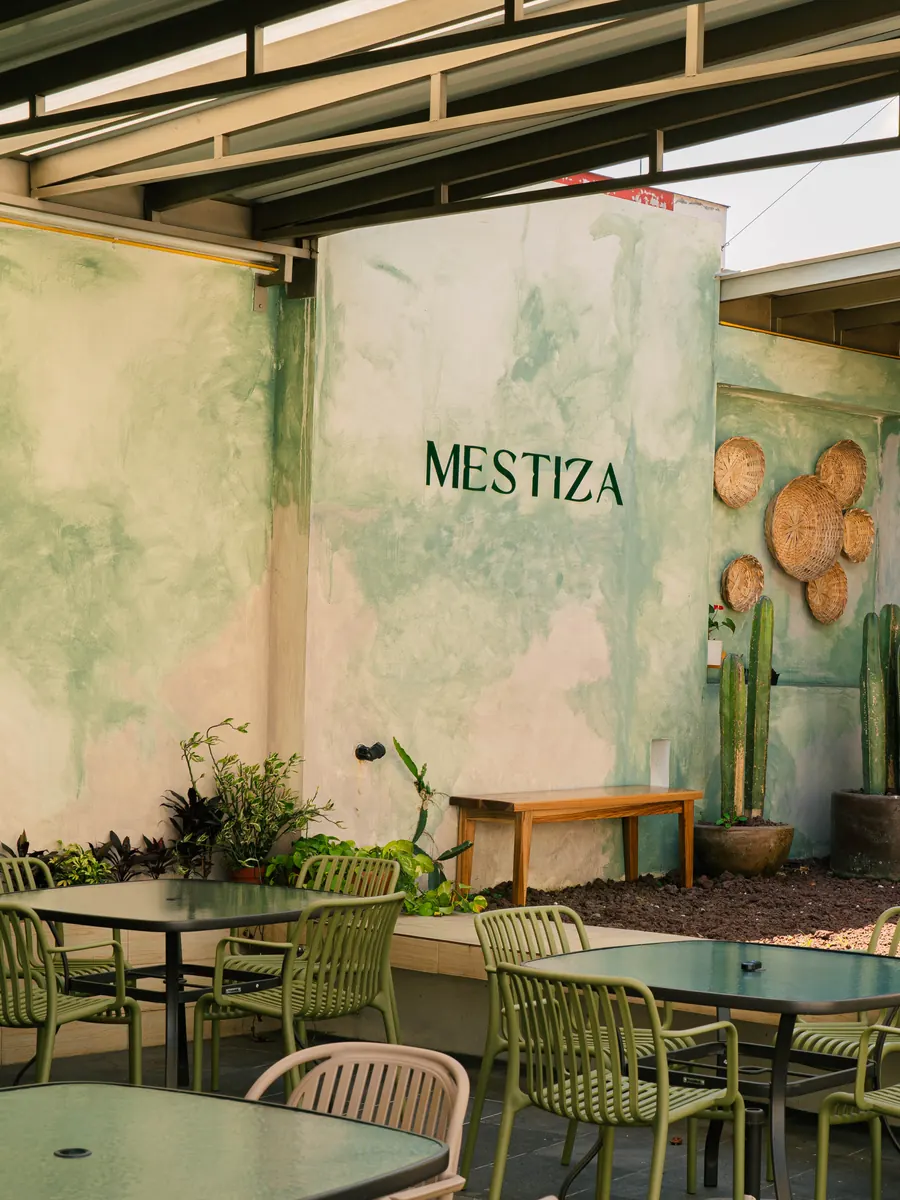 Patio de Mestiza: muro de estuco verde salvia con el nombre del restaurante pintado, tenates de palma colgados, cactus en macetas de barro y mesas del comedor al aire libre